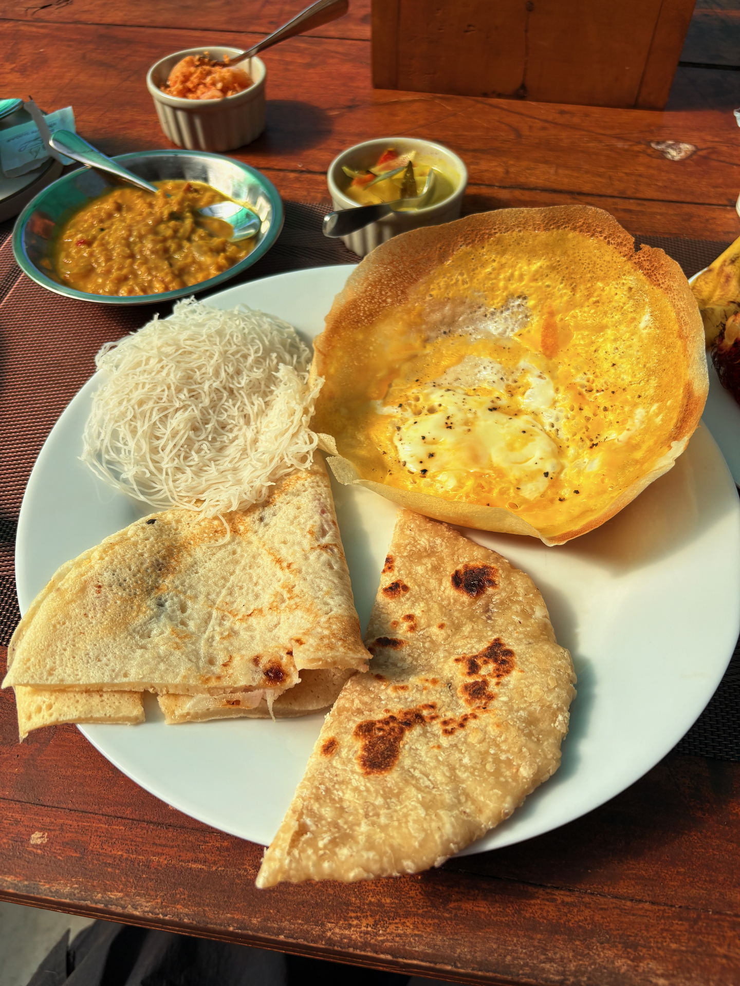 A plate of egg hoppers and rice with side dishes on a timber table.