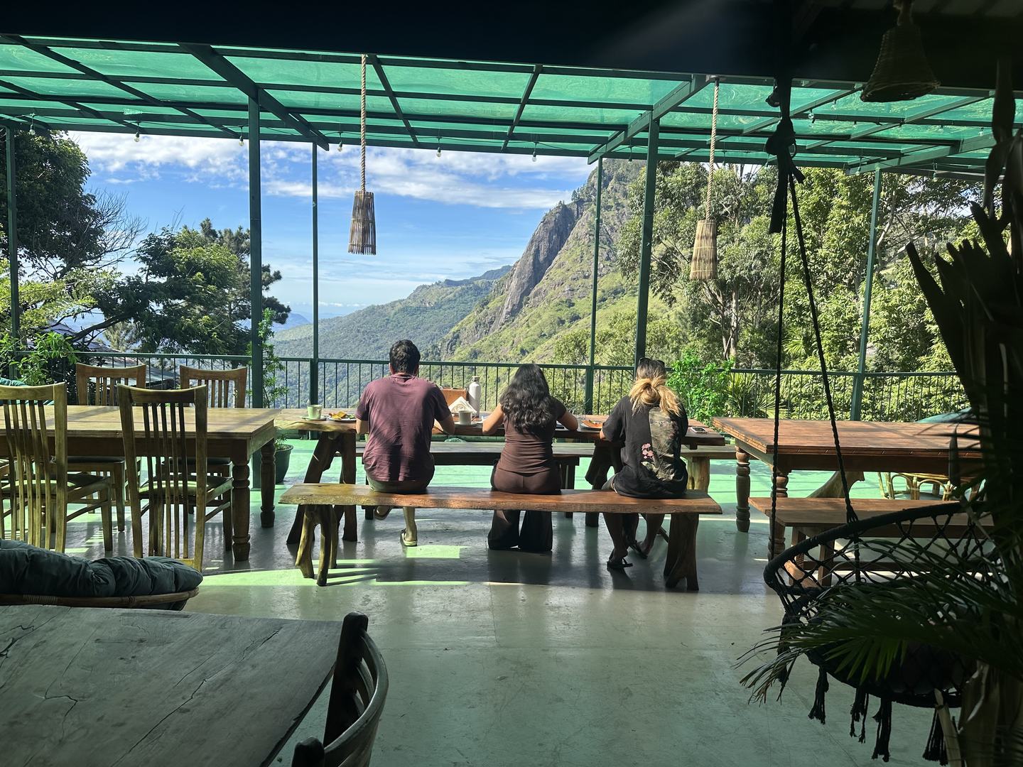 Three guests sitting side by side on a timber bench at breakfast, seen from behind, facing the railing and the rock face across the valley.