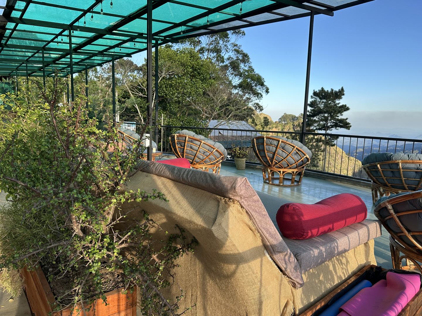 An empty cushioned daybed and round rattan chairs on the terrace under the green roof, with the valley beyond the railing.