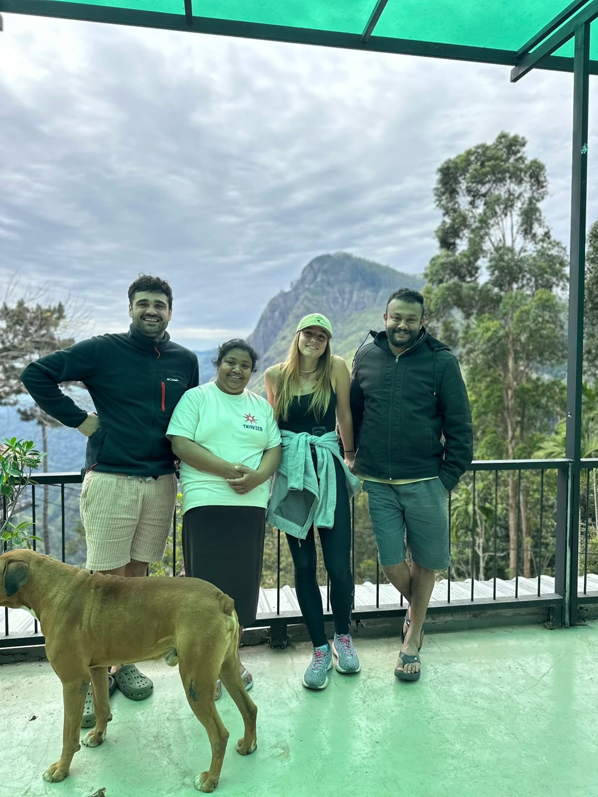 Four people standing at the terrace railing under the green roof, the ridge across the valley behind them and Sando the dog in the foreground. Sithara Wimalasooriya is on the right.