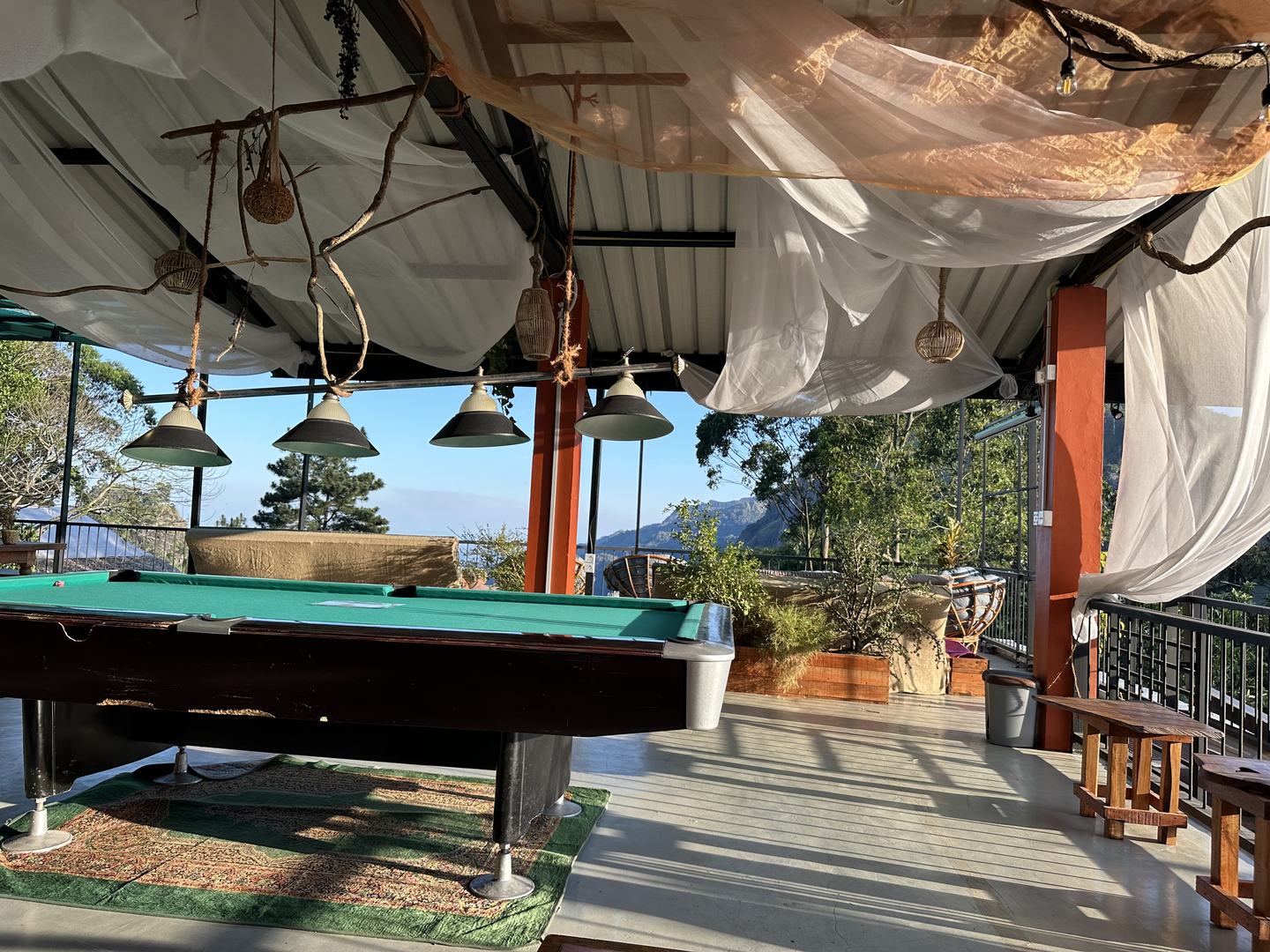 A pool table on the terrace under four hanging metal lamps, with the railing and a mountain ridge beyond it.